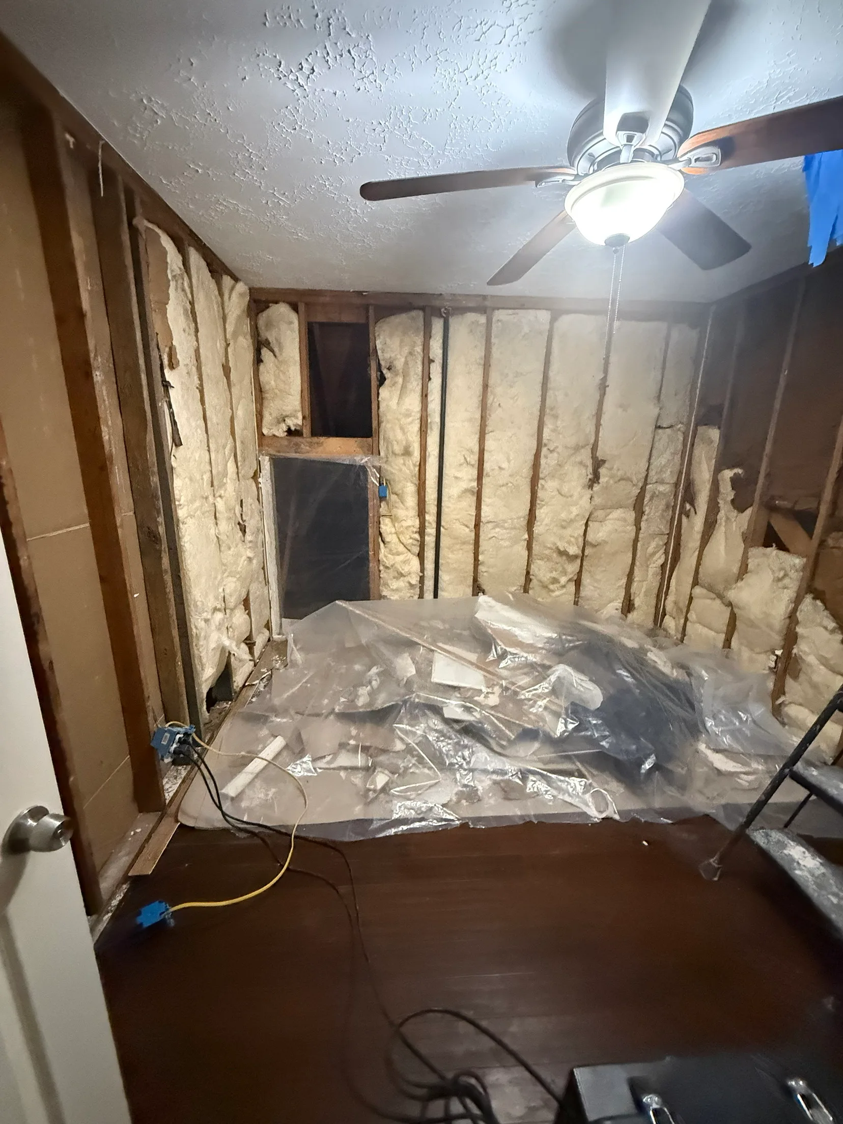 A bedroom stripped back to the studs with insulation exposed on every wall and debris gathered under plastic sheeting on the floor.