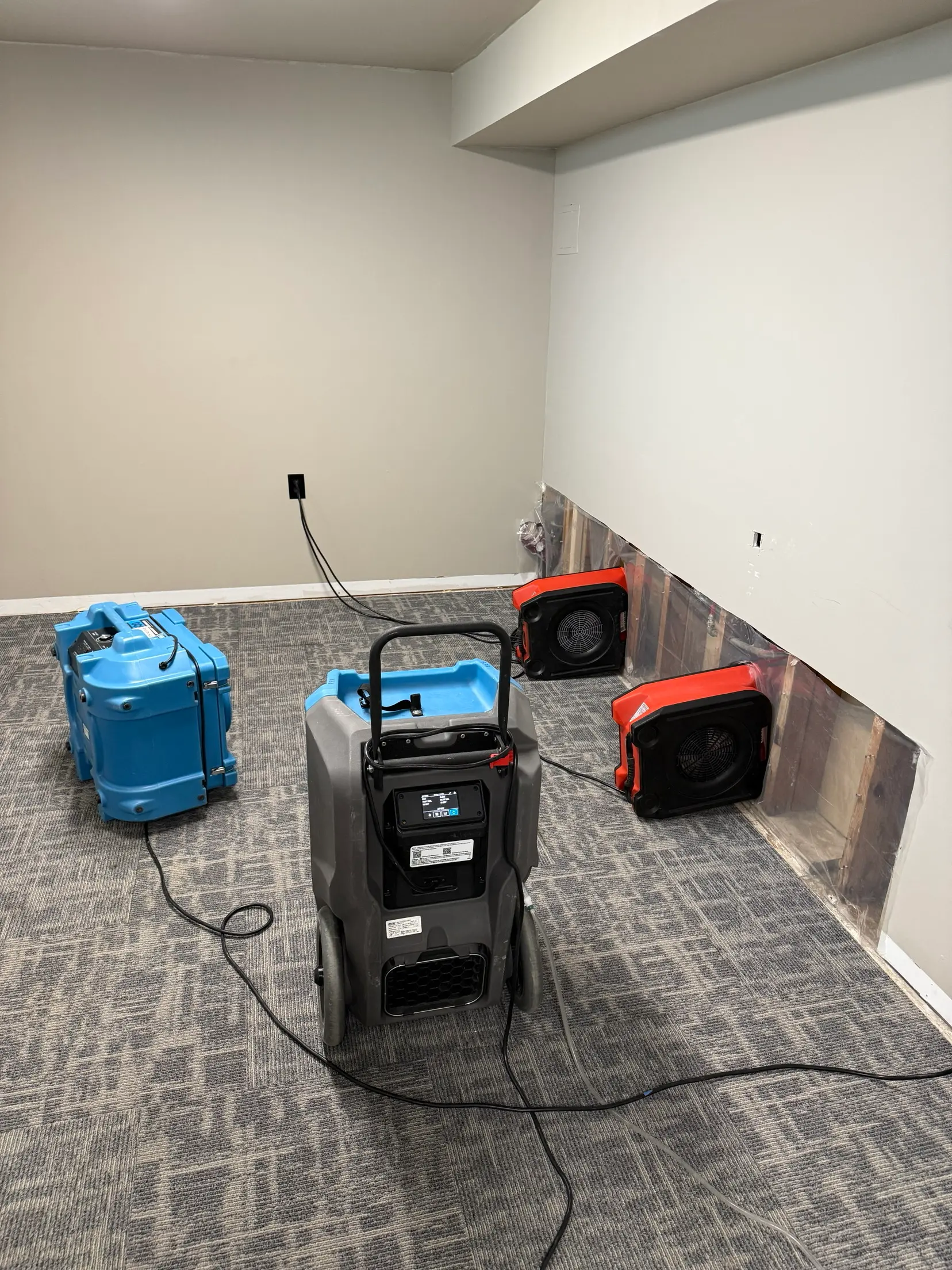 Structural drying in progress: a blue air scrubber, an LGR dehumidifier and two orange axial air movers directed into an open wall cavity where the baseboard has been removed and the wall lined with plastic sheeting.