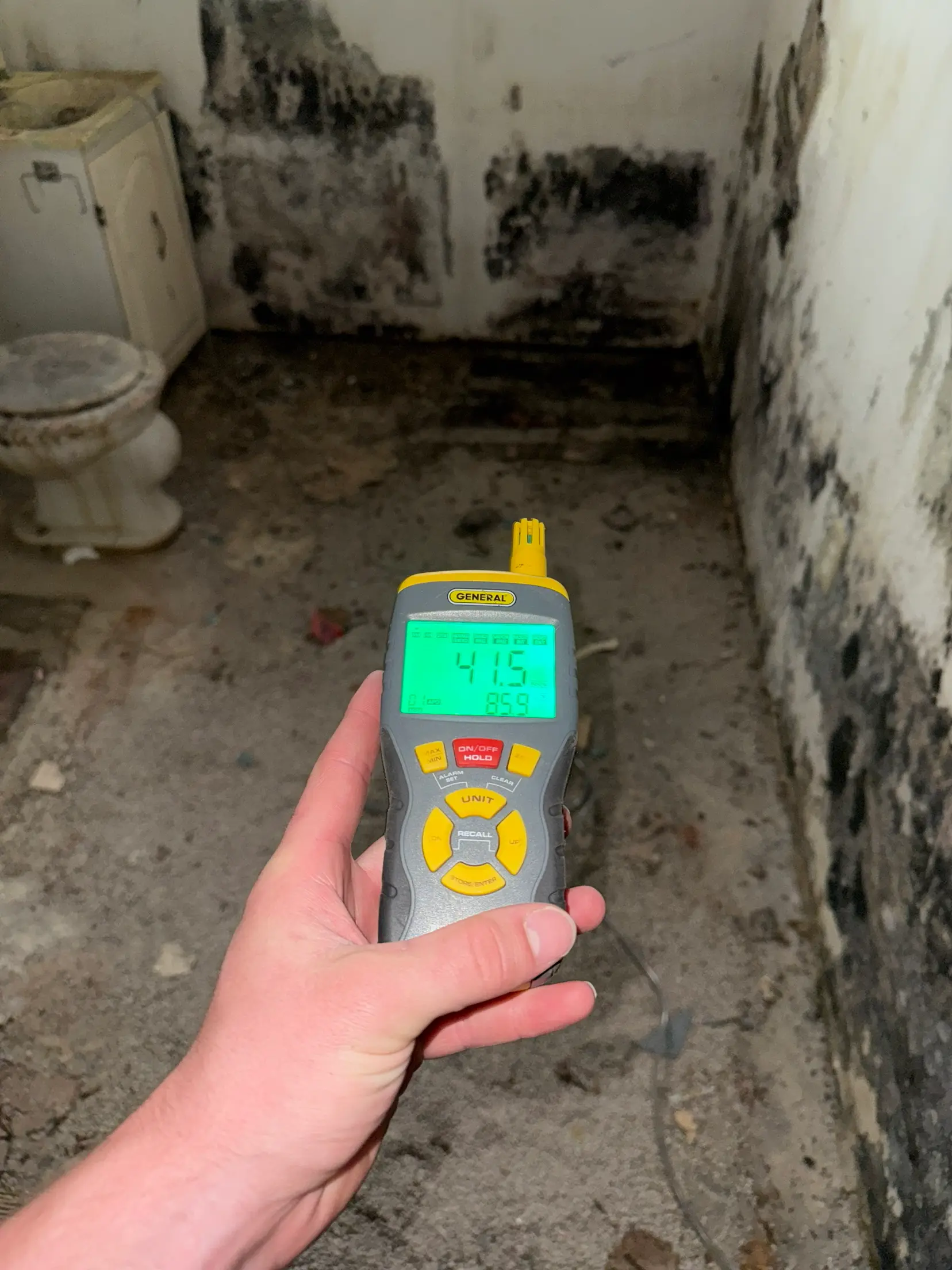 A thermo-hygrometer reading 41.5 percent relative humidity at 85.9 degrees, held up in a bathroom where black mold covers the lower walls and the floor has been stripped back to the slab.