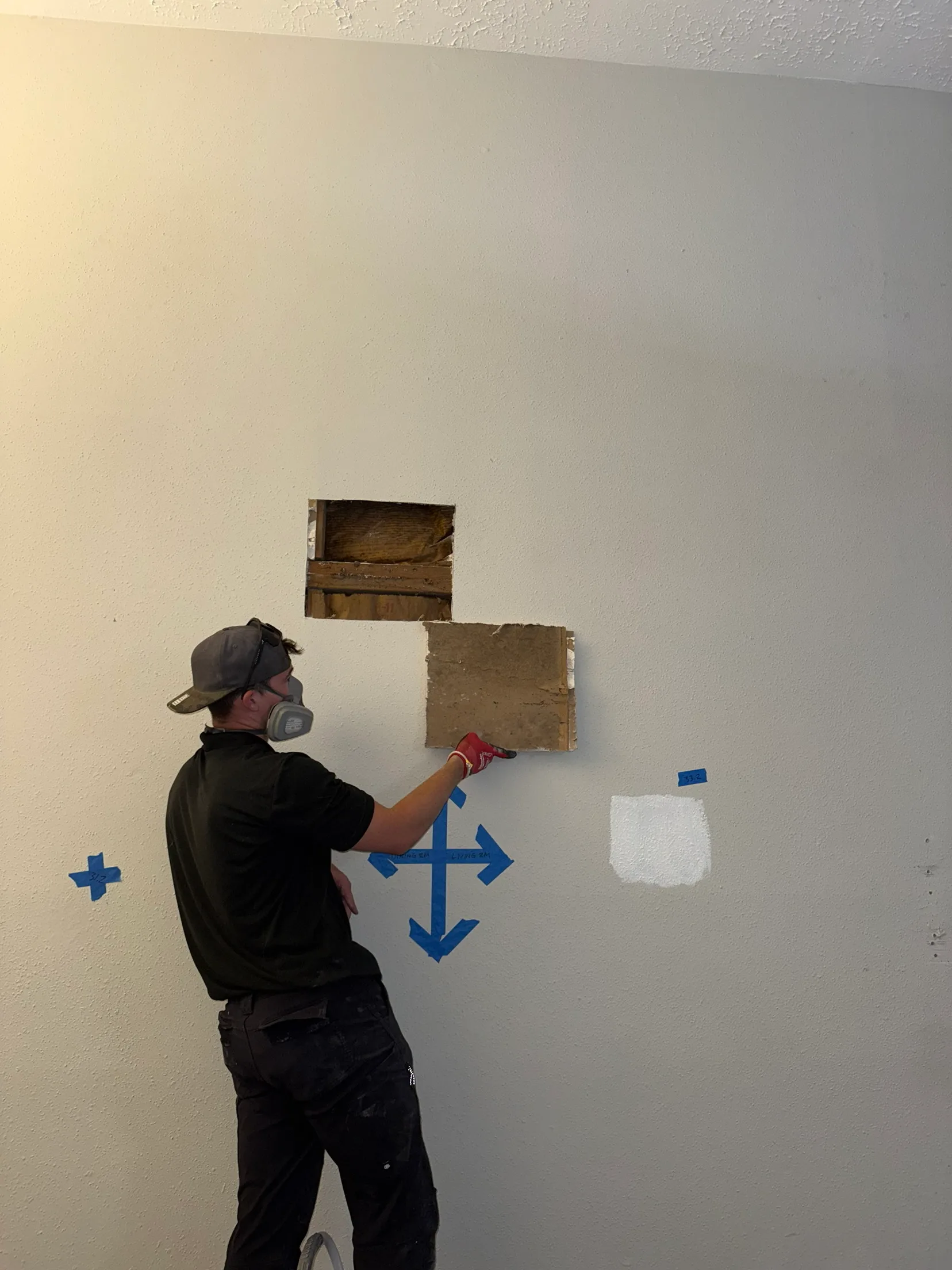 An Elite Home Industries technician wearing a half-face respirator, holding a section of drywall cut out of a wall for inspection, with blue tape arrows on the wall marking room boundaries.